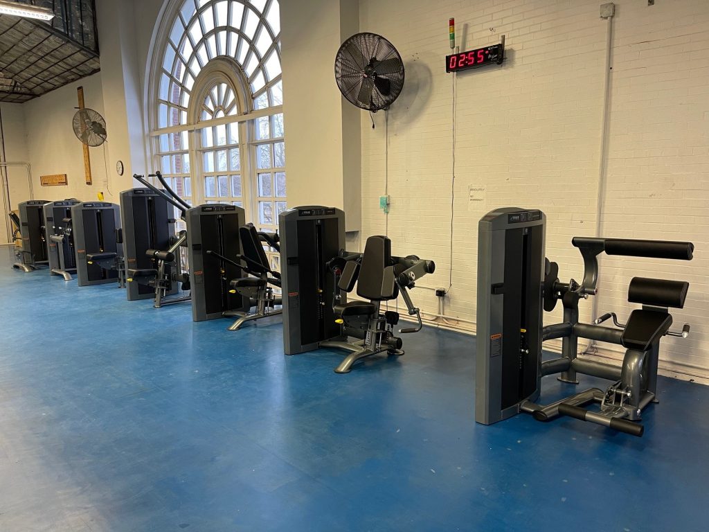 Row of black gym machines on a blue floor next to a white brick wall with large arched windows, wall fans, and a digital clock displaying 02:55. Room is empty and well-lit.