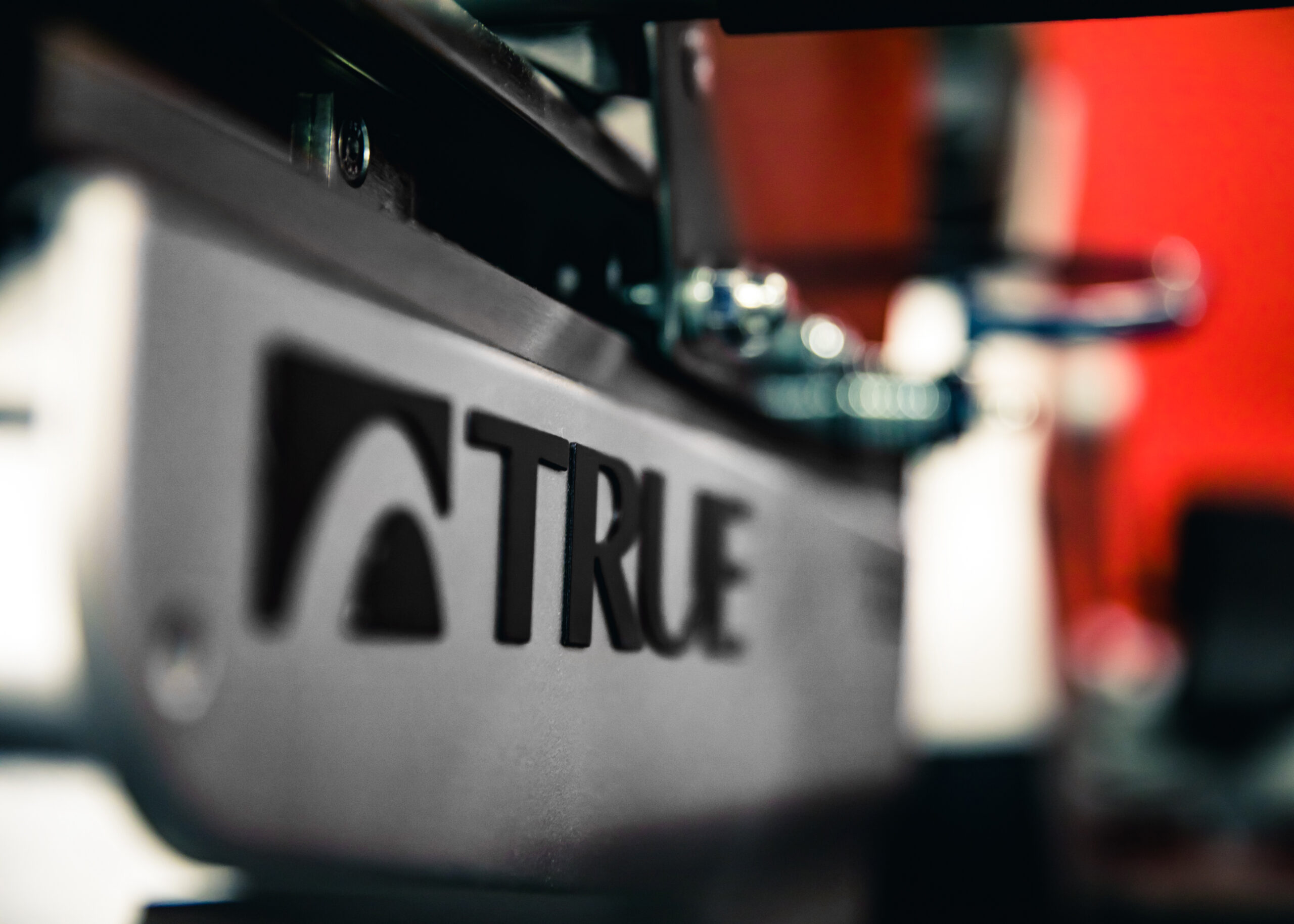 Close-up of a metallic surface with the word TRUE and a logo engraved on it; background is blurred with hints of red and blue tones.