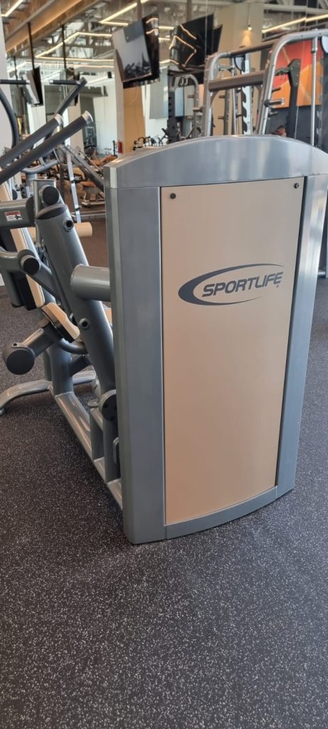 A Sportlife-branded gym machine with padded handles and seats stands on a speckled gym floor, surrounded by other exercise equipment in a fitness center.