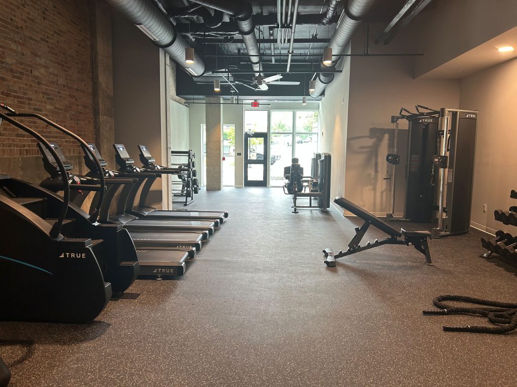 A gym with treadmills, ellipticals, weight machines, free weights, and a bench. There are large windows and a door at the far end, with exposed ducts on the ceiling and brick walls on the left side.