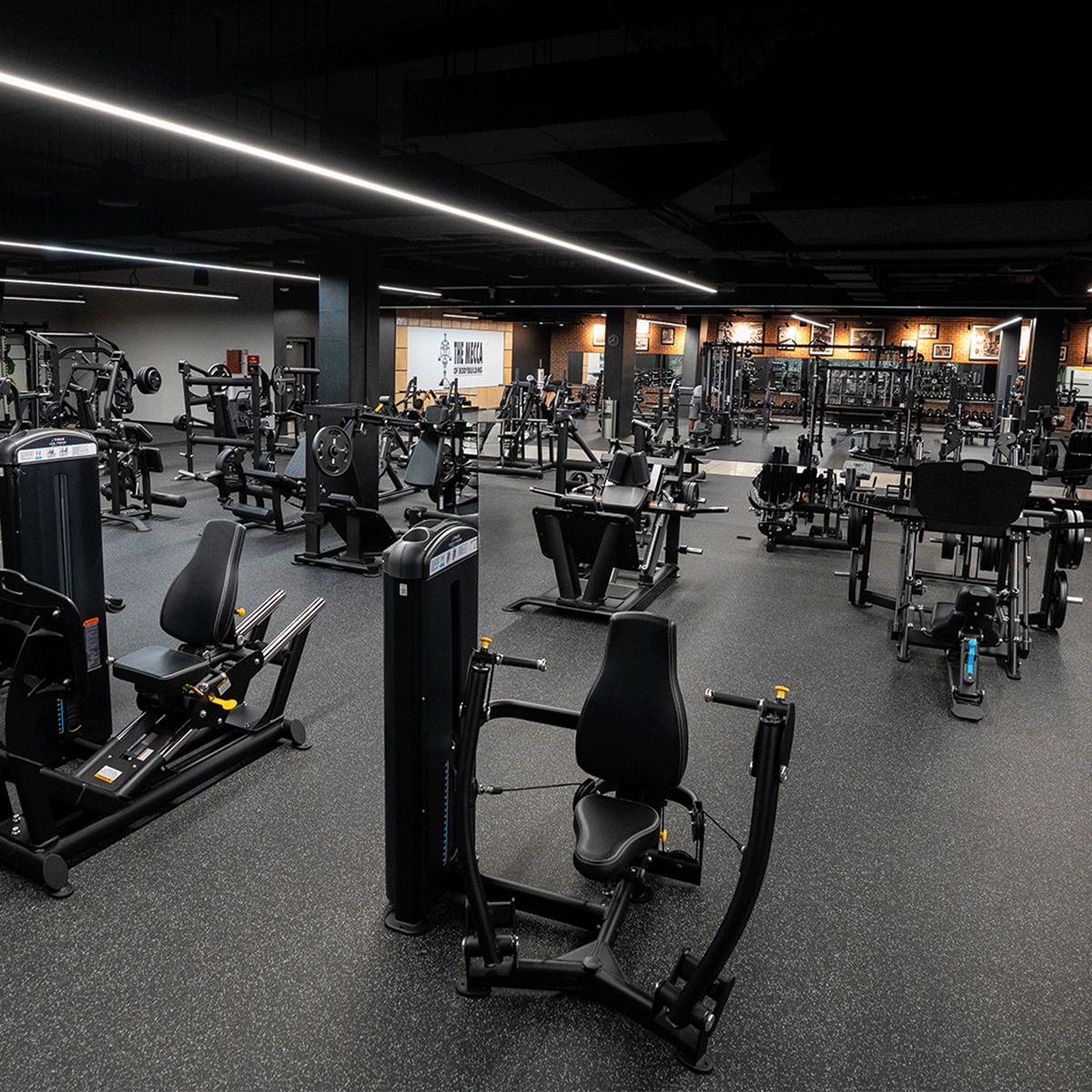 A spacious, modern gym with various strength training machines and equipment arranged neatly on a clean, black floor. The gym is well-lit and empty, with no people present.