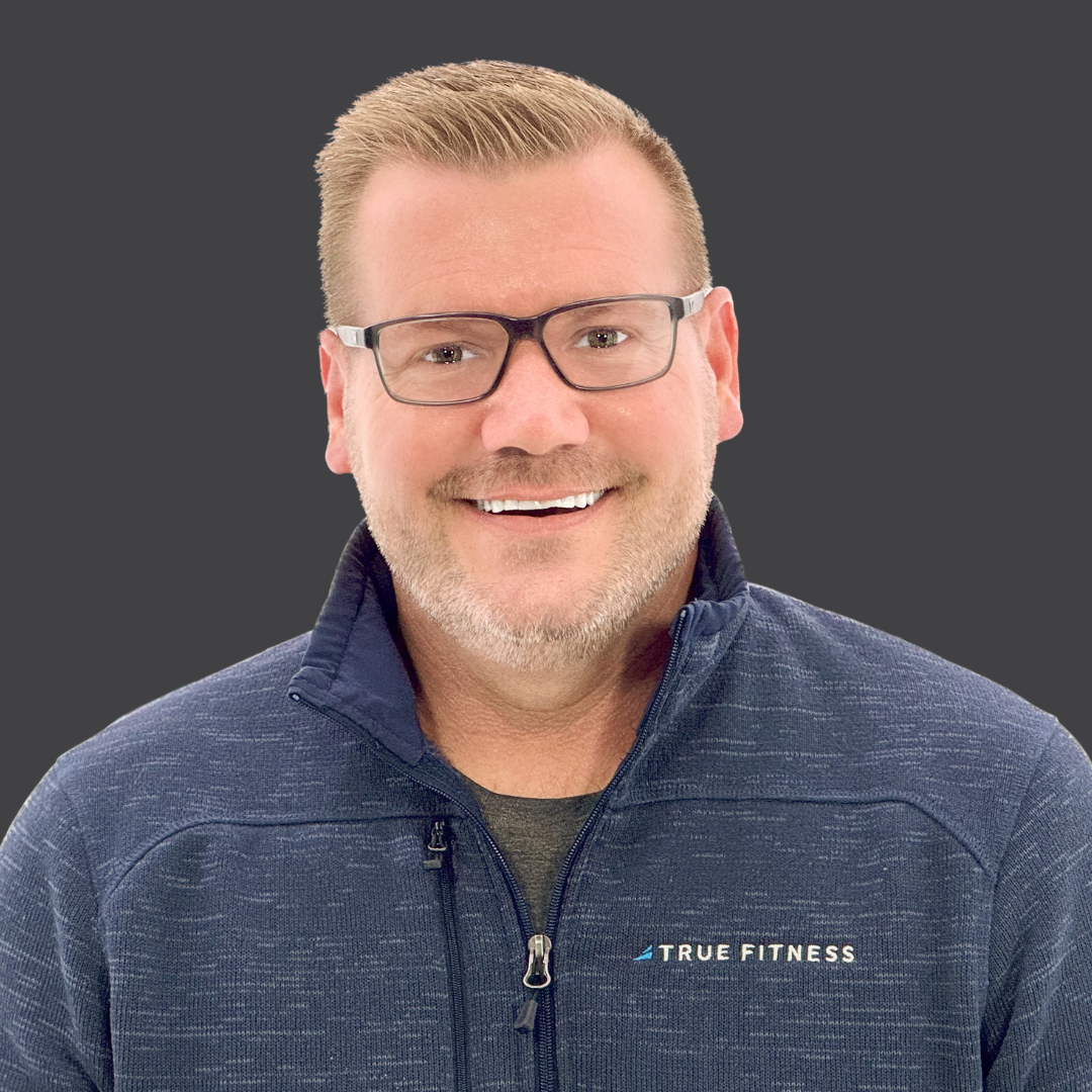 A man with short light hair, glasses, and a trimmed beard is smiling. He is wearing a navy zip-up jacket with the words 66体育 - TRUE Fitness让运动更简单_官方网站 66体育 66体育 on it, and the background is a solid dark gray.