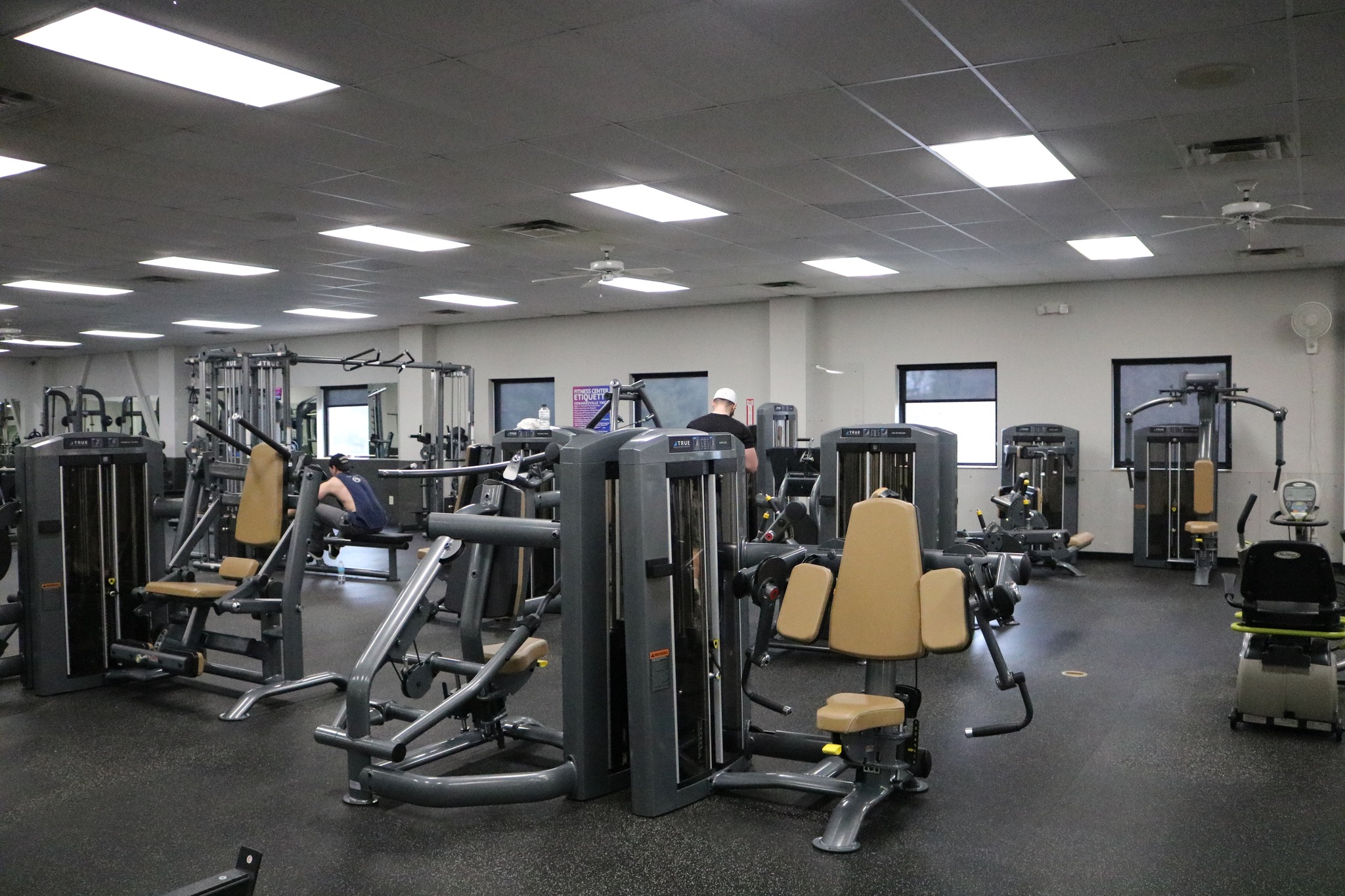 A gym with various exercise machines, including weight and cardio equipment, arranged in rows. The room has overhead fluorescent lighting, beige and gray machines, and a few people working out in the background.