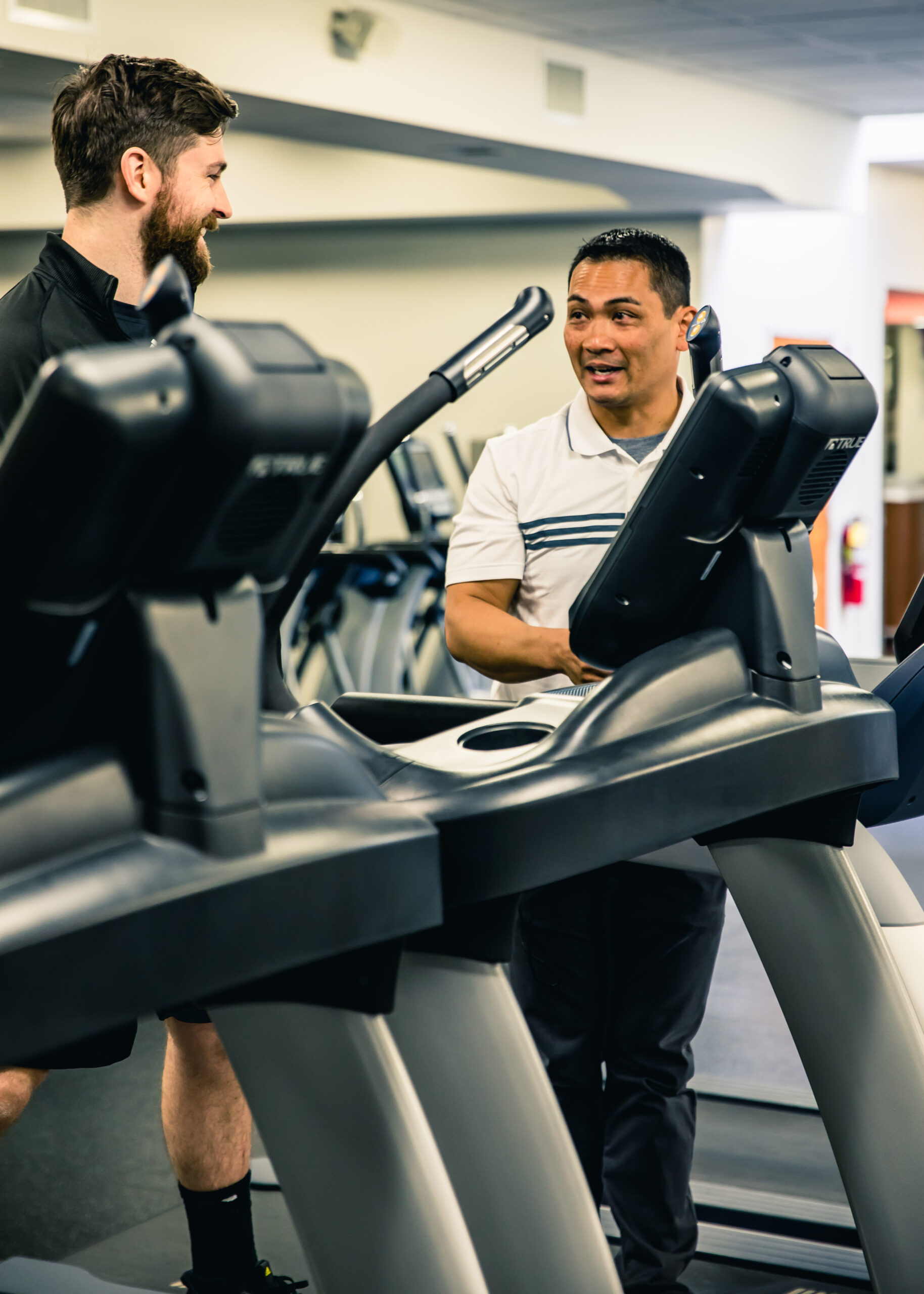 Two men are standing next to treadmills in a gym, engaged in conversation. One man is wearing a black outfit, while the other wears a white shirt with stripes. Exercise equipment is visible in the background.