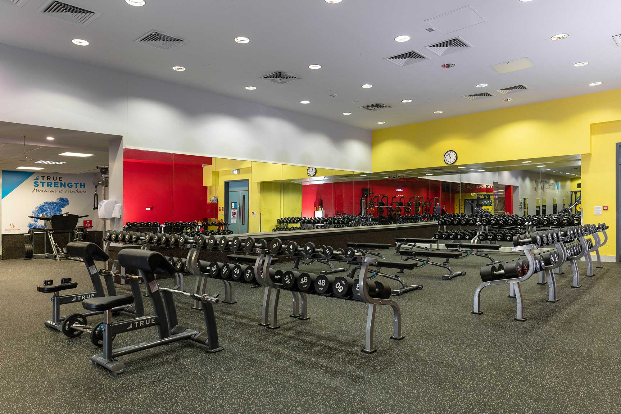 A spacious gym with rows of dumbbells, weight benches, and strength training equipment. Large mirrors cover the yellow and red walls, and a clock is mounted above the mirrors. The floor is covered with dark rubber matting.