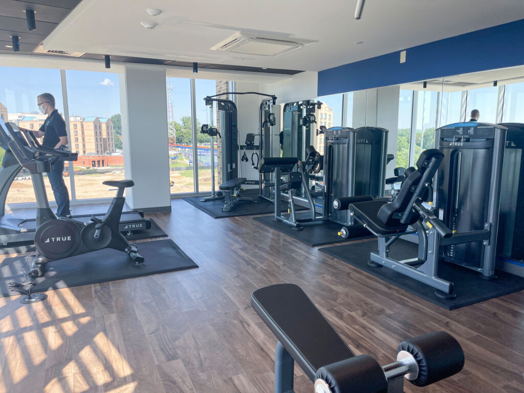 Modern gym with large windows, various strength training machines, a stationary bike, bench, and dumbbell. One person is using the treadmill near the windows with a cityscape visible outside.