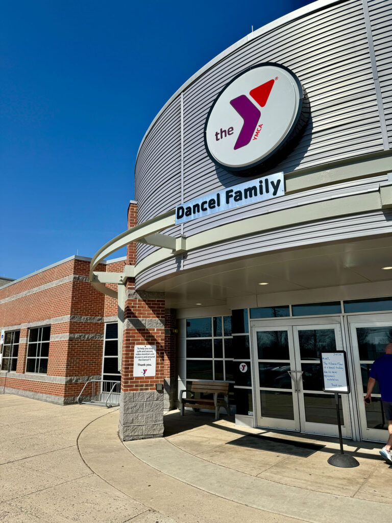 Entrance of a YMCA building labeled Dancel Family with a large YMCA logo above the doors, brick and metal exterior, clear sky, and signs near the entrance.
