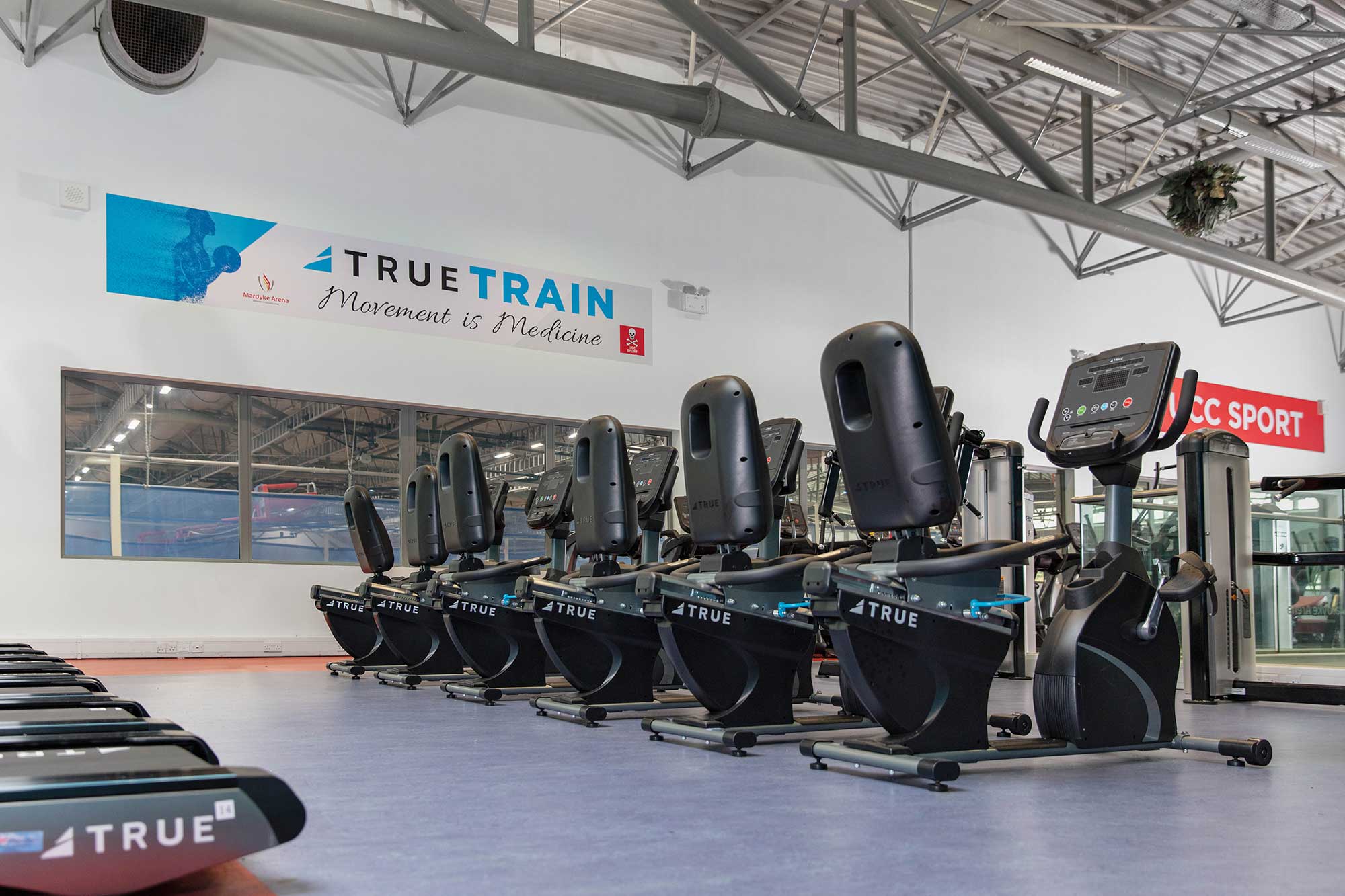Row of recumbent exercise bikes and treadmills in a spacious, modern gym with high ceilings; large sign on the wall reads TRUE TRAIN Movement is Medicine.
