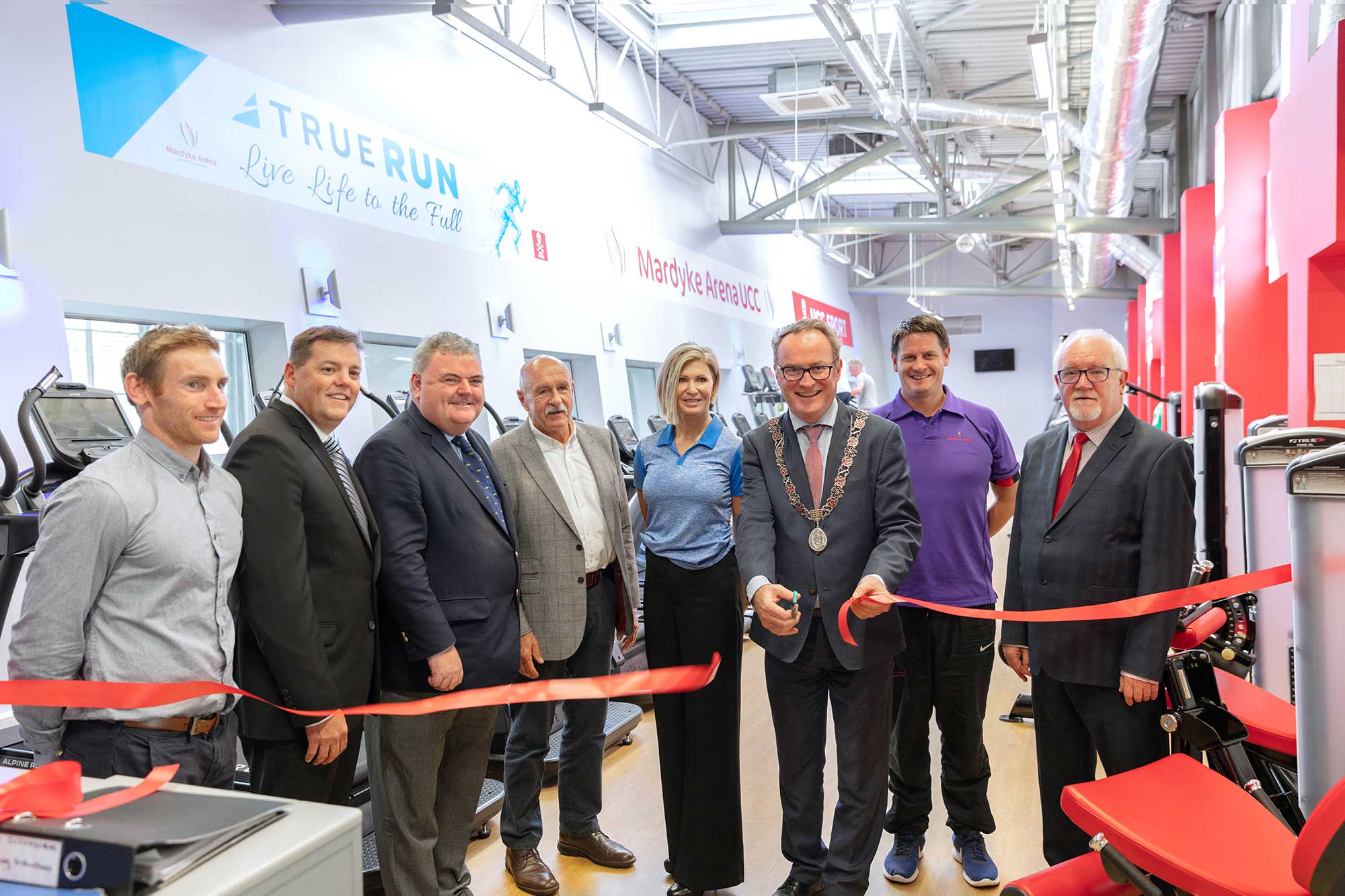 A group of people stand in a gym for a ribbon-cutting ceremony. Several men wear suits, while others are in casual clothes. One man, wearing a ceremonial chain, holds scissors and smiles as he cuts the red ribbon. Gym equipment is visible behind them.
