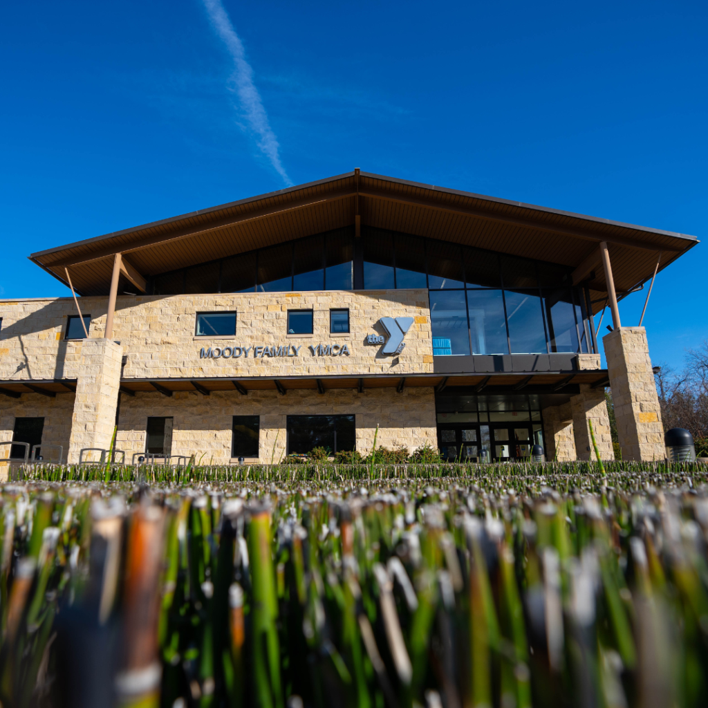 Low-angle view of the Moody Family YMCA building with tan stone walls, large windows, and a sloped roof, against a clear blue sky. Trimmed green grass is visible in the foreground.