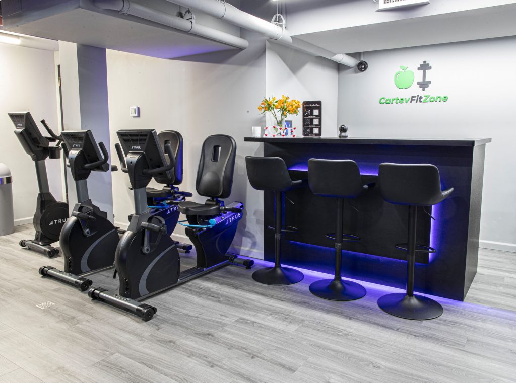 A modern gym area with three stationary exercise bikes next to a sleek black bar counter with four stools; the wall displays CarteyFitZone and a green apple logo, with flowers and a coffee machine on the counter.