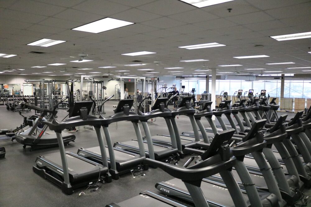 A spacious, modern gym with rows of treadmills in the foreground and various strength training machines and equipment in the background, well-lit by overhead lights and large windows.