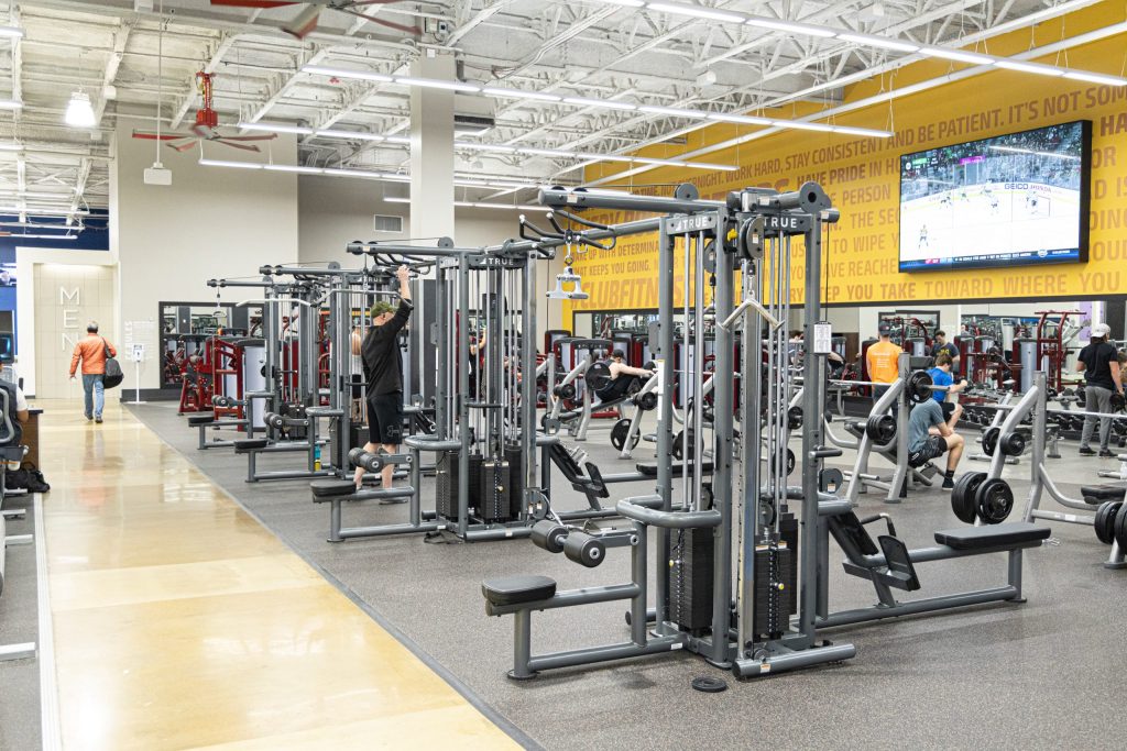 A spacious gym with various exercise machines and people working out. The walls are decorated with motivational quotes, and a large TV displays a basketball game. The gym has bright lighting and a clean, organized layout.