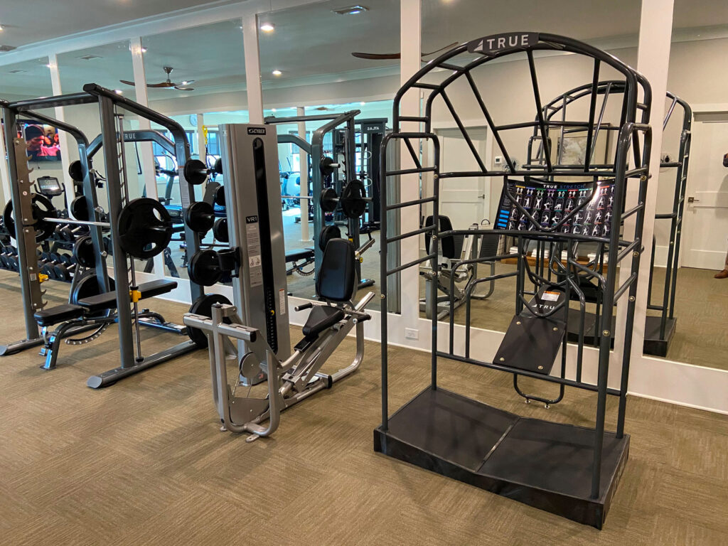 A gym with various equipment including weight machines, free weights on racks, and a TRUE stretch cage, with mirrors covering the back wall and beige carpet flooring.