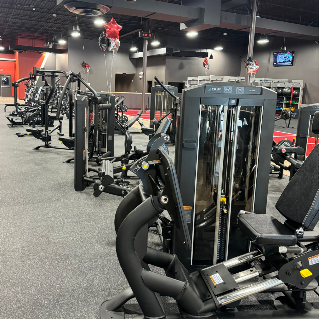 A modern gym with various exercise machines, black flooring, and red star balloons hanging from the ceiling. The space is well-lit and spacious, with strength training equipment arranged neatly.