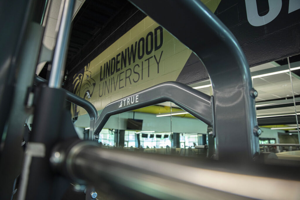 Close-up of gym equipment with a metal bar, seen in front of a wall displaying Lindenwood University and a lion logo in a fitness center. Fluorescent lights and mirrors reflect the room.