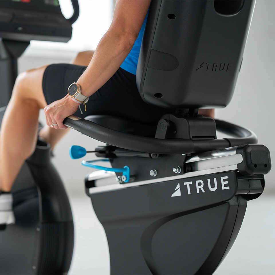 A person wearing a blue top and black shorts exercises on a recumbent stationary bike by TRUE in a bright indoor space. The person’s hand is resting on the side handlebar.