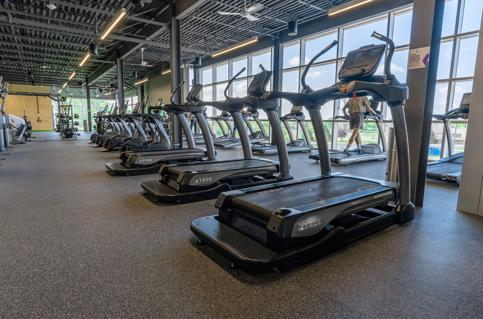 A row of treadmills in a modern gym with large windows letting in natural light; one person is using a treadmill in the background. The gym has black flooring and an industrial-style ceiling.
