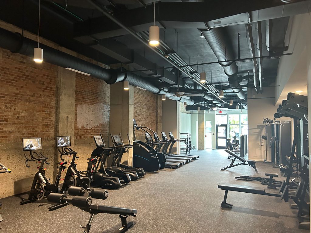 A modern gym with exposed brick walls and industrial ceilings, featuring stationary bikes, treadmills, ellipticals, weight benches, and various exercise equipment, with natural light coming from large front windows.
