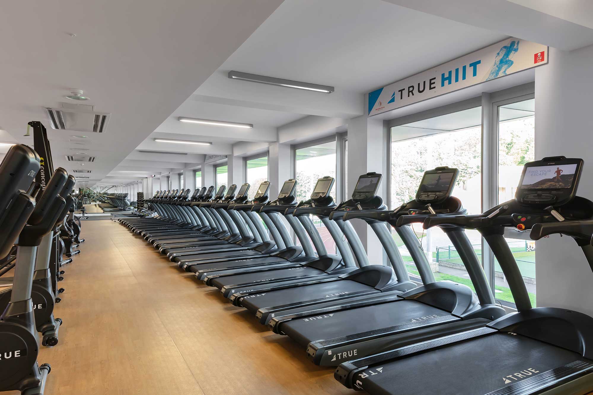 Rows of treadmills are lined up in a bright, modern gym with large windows. The room is empty, and a “TRUE HIIT” sign is visible above the windows, letting in natural light.