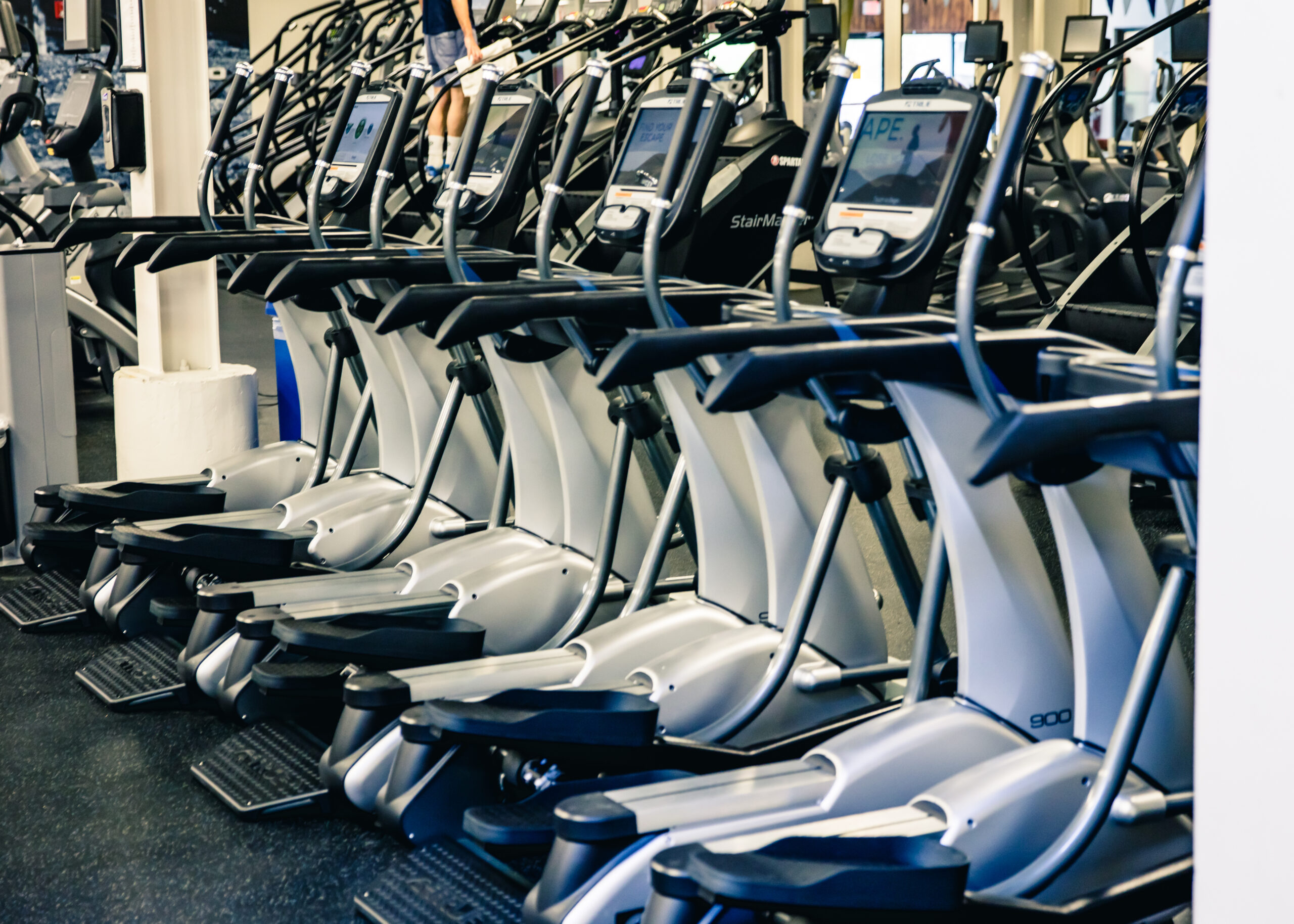 A row of elliptical machines with digital screens lined up in a gym, ready for use. The gym has a clean, modern appearance with other fitness equipment visible in the background.
