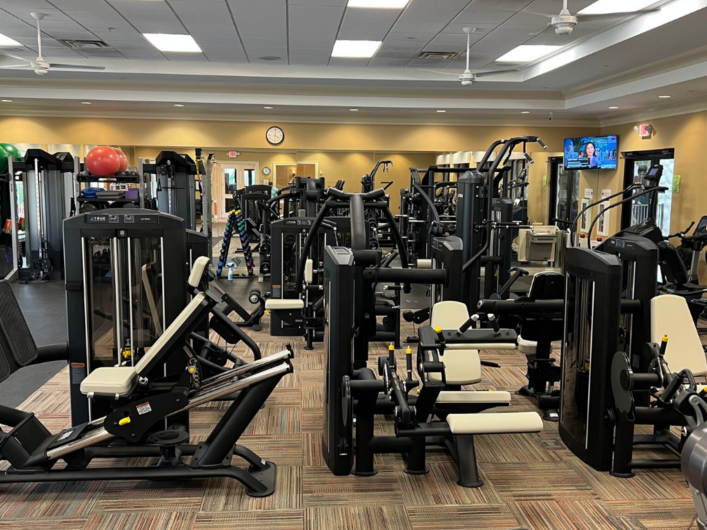 A spacious gym with various weight machines, exercise equipment, and free weights. The room has beige walls, ceiling fans, a wall clock, a TV, and ample lighting with no people present.
