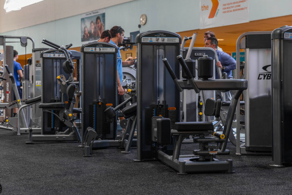 Rows of strength training machines in a gym, with several people working out in the background. The gym has dark flooring and exercise equipment arranged in neat lines.