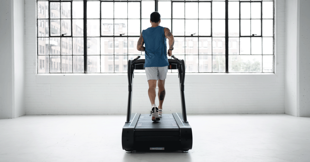 A man in athletic wear runs on a treadmill in a bright, spacious gym with large industrial-style windows and a cityscape visible outside.