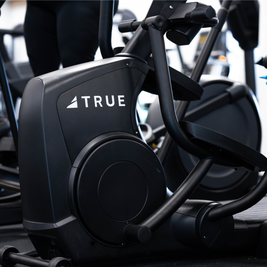 A close-up of a black elliptical exercise machine labeled TRUE in white letters, located in a gym with other fitness equipment visible in the background.