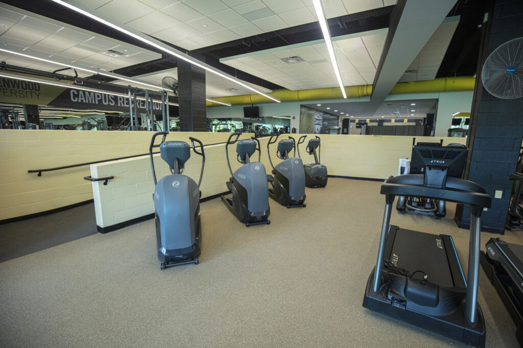 A gym room with four elliptical machines lined up side by side and two treadmills nearby; mirrors cover part of the walls and overhead fluorescent lights illuminate the space.
