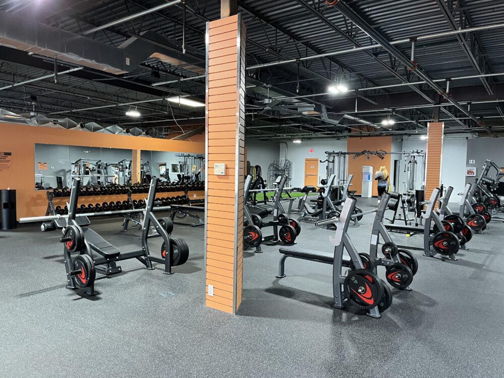 A spacious gym with various weightlifting equipment, including benches, barbells with red-accented weights, and dumbbells. Large mirrors line the back wall; a person is visible near the rear exercise machines.