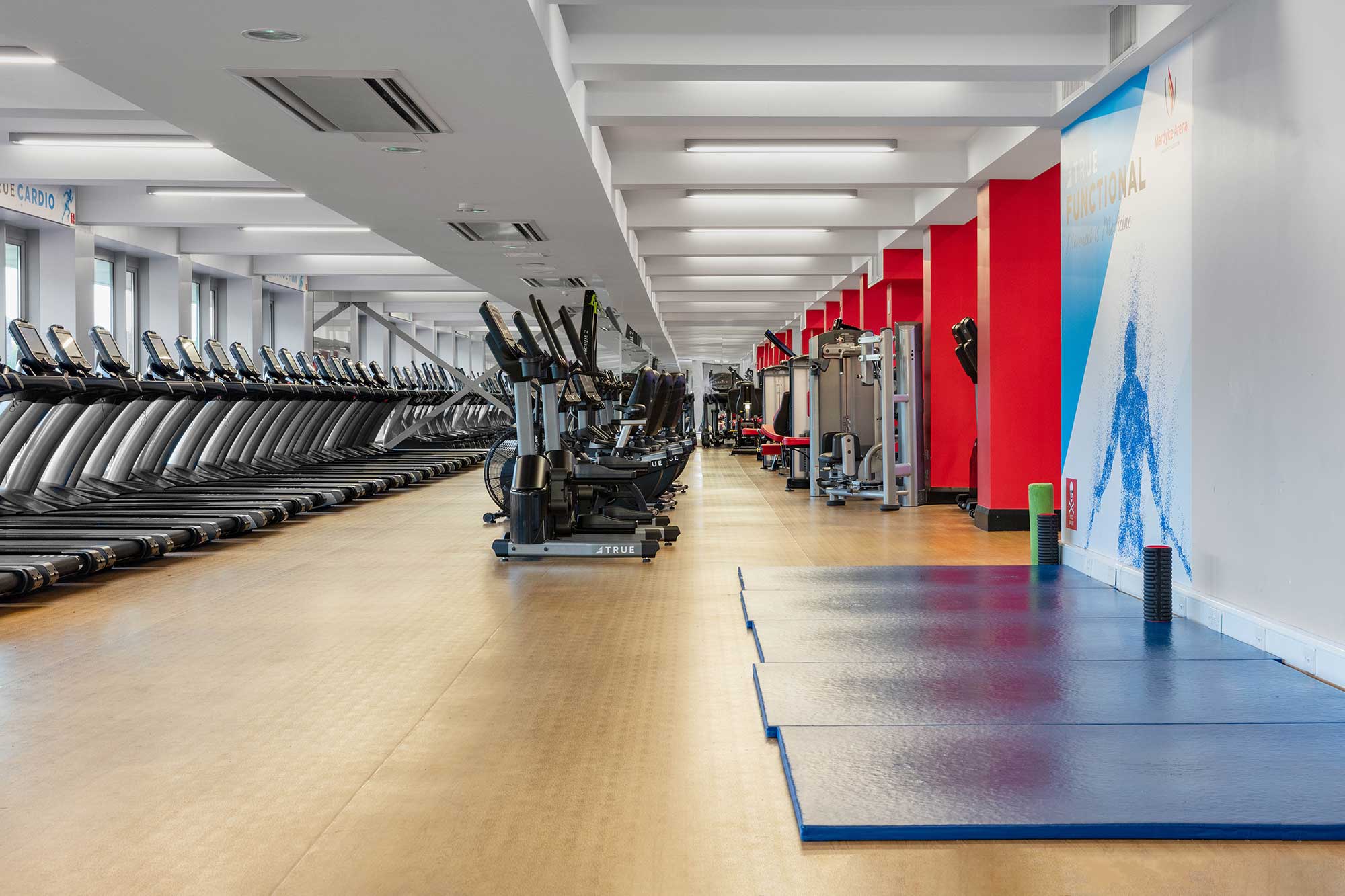 A spacious, modern gym with rows of treadmills and elliptical machines on the left, weight machines in the center, and blue exercise mats on the right, all lit by overhead lights and large windows.