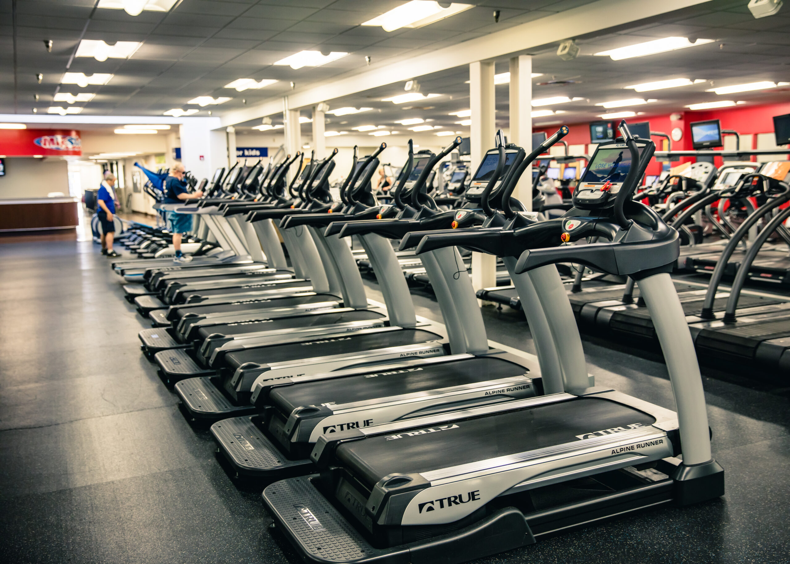 A row of treadmills lines the floor in a spacious, well-lit gym with various other fitness equipment visible in the background. A few people are using the machines or walking around the gym area.