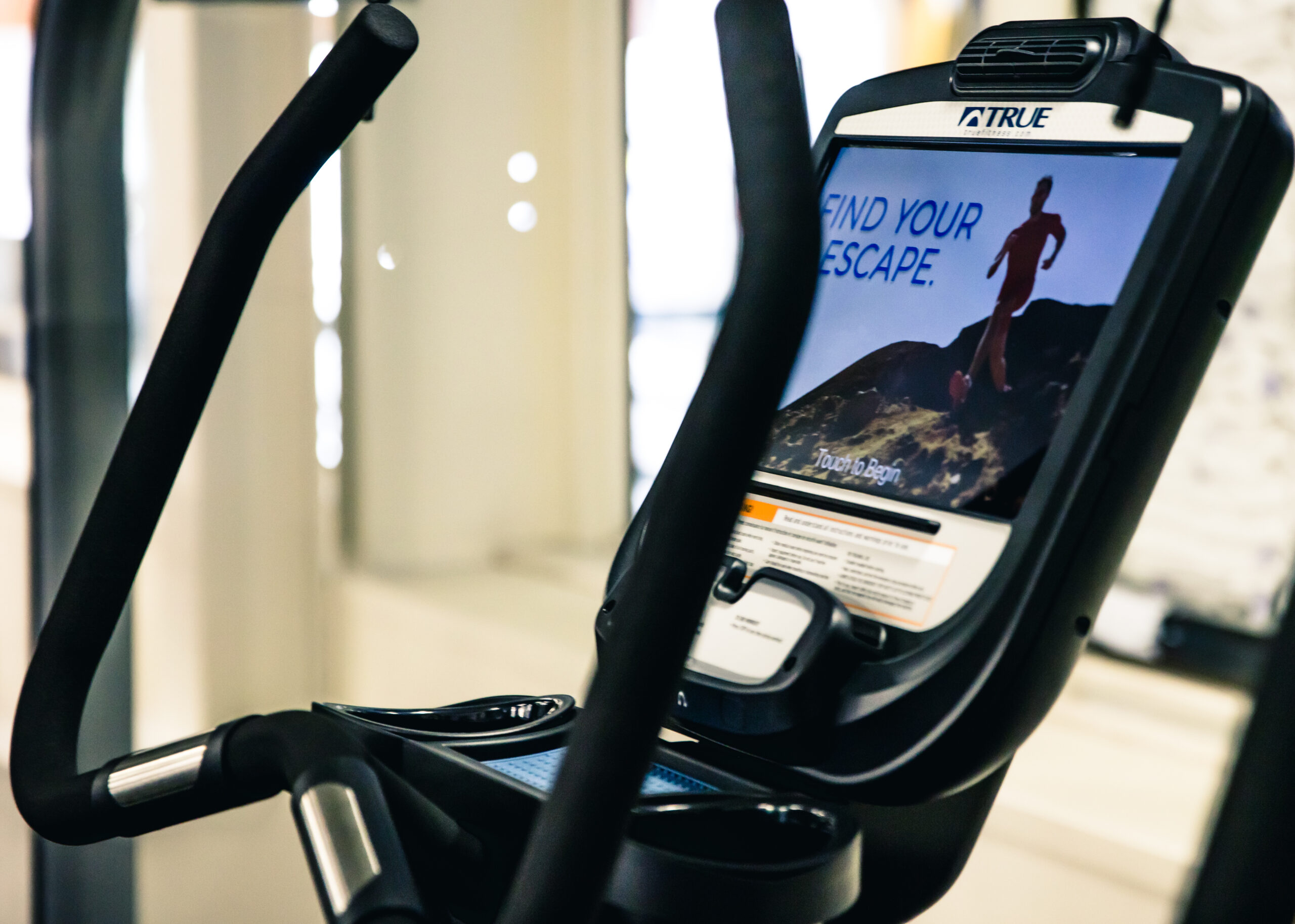 Close-up of an elliptical machine’s display screen showing an image of a person running outdoors with the words “FIND YOUR ESCAPE.” The exercise machine’s handlebars and control panel are also visible.