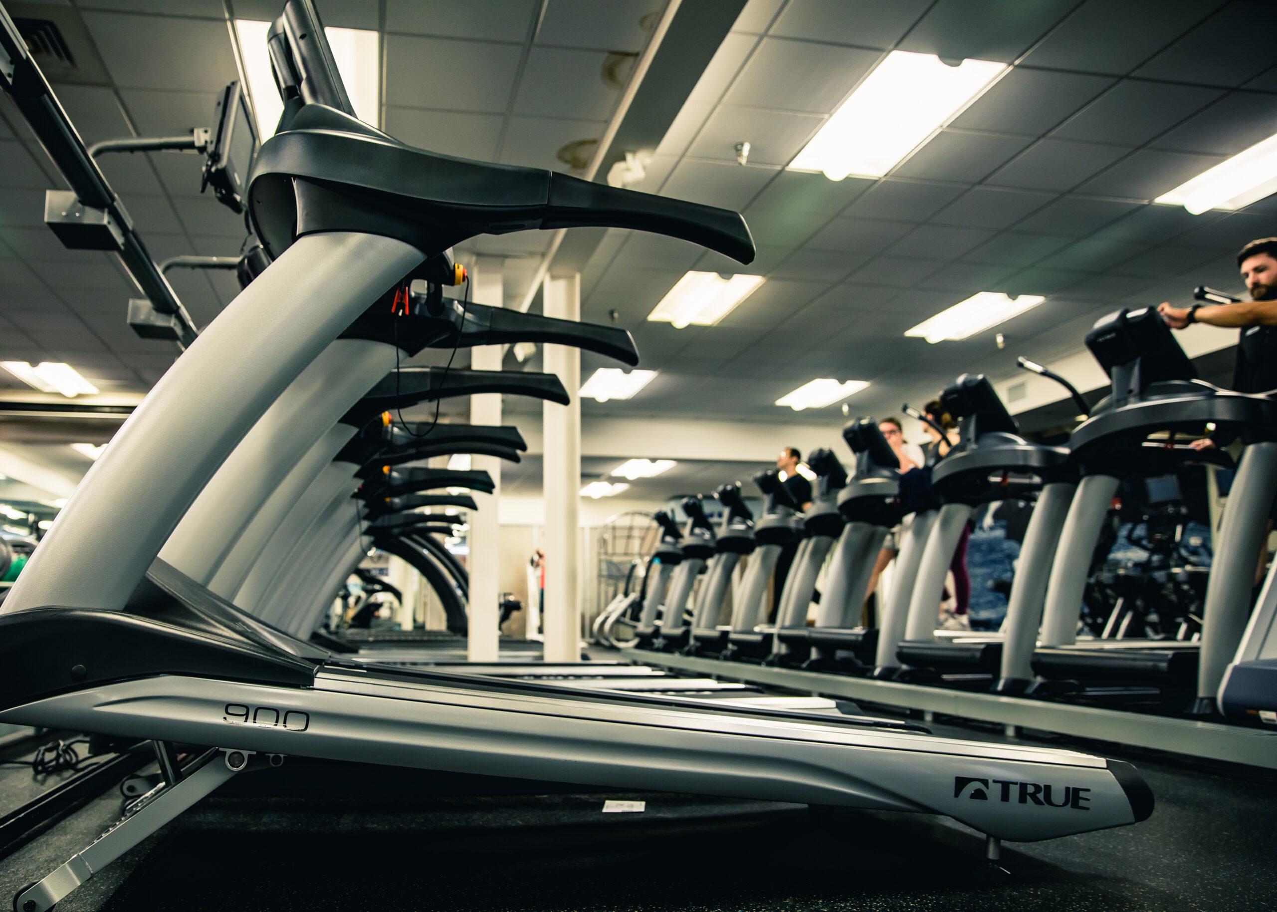 Row of treadmills in a gym, viewed from a low angle. Several people are walking or running on the treadmills in the background under bright ceiling lights.