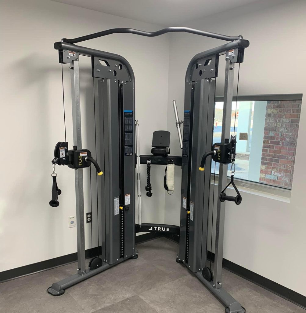 A cable crossover machine with adjustable pulleys and handles stands in the corner of a gym room with gray flooring and white walls, next to a window.