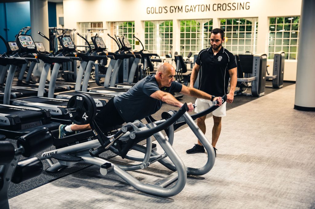A man works out on a Hammer Strength machine while a trainer observes and guides him in a gym with treadmills in the background. The sign reads Golds Gym Gayton Crossing.