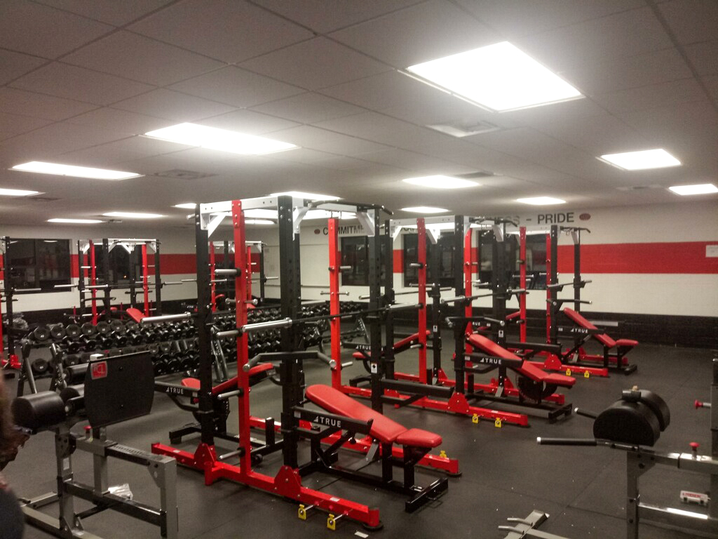 A gym room with red and black weightlifting equipment, benches, barbells, squat racks, and mirrors along the walls. The room has bright overhead lighting and a black and red accent wall with the word PRIDE displayed.