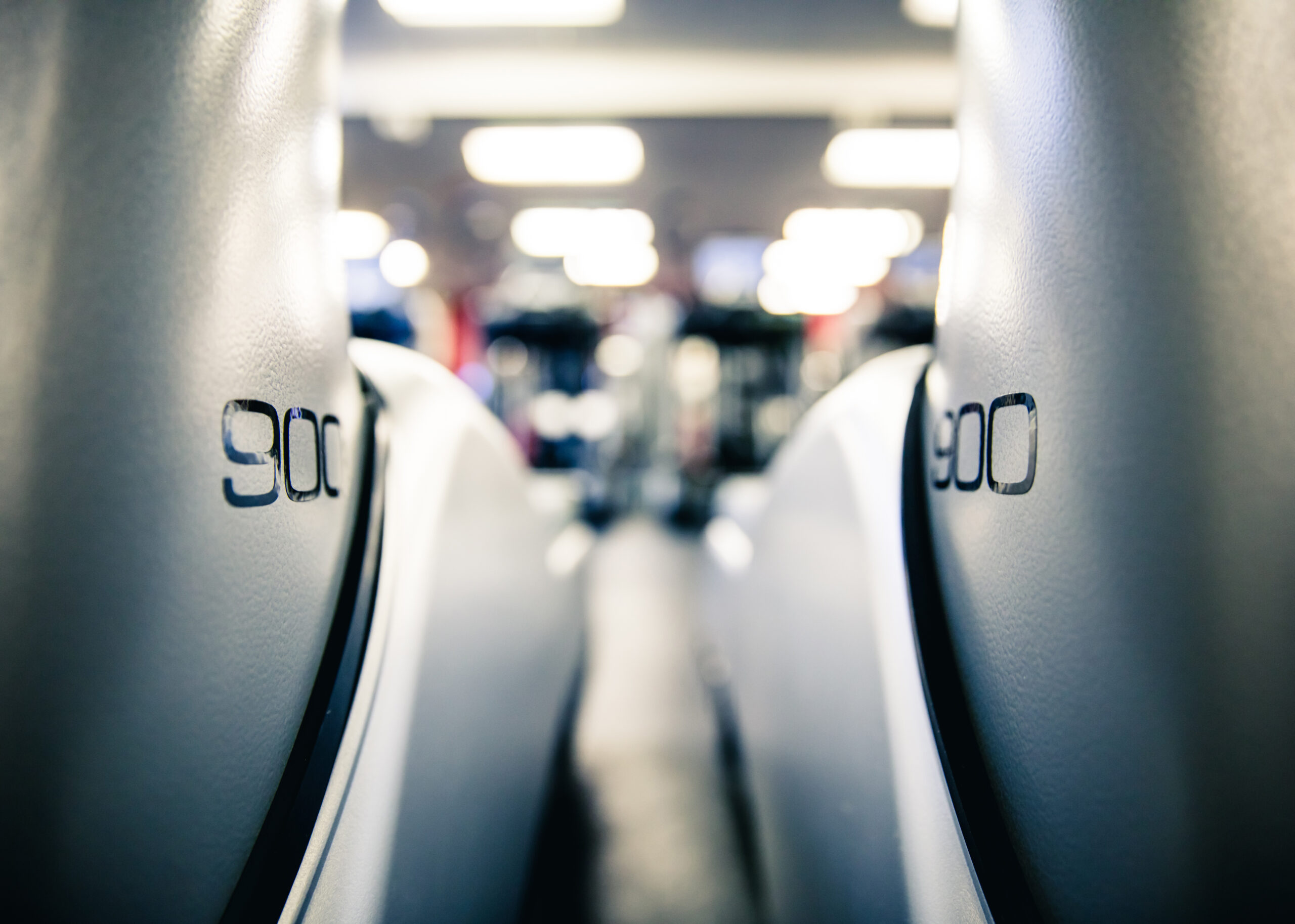 Close-up view of two gray seats with 900 printed on the backrests, facing toward a brightly lit, blurred interior space, possibly in a gym or public transportation setting.