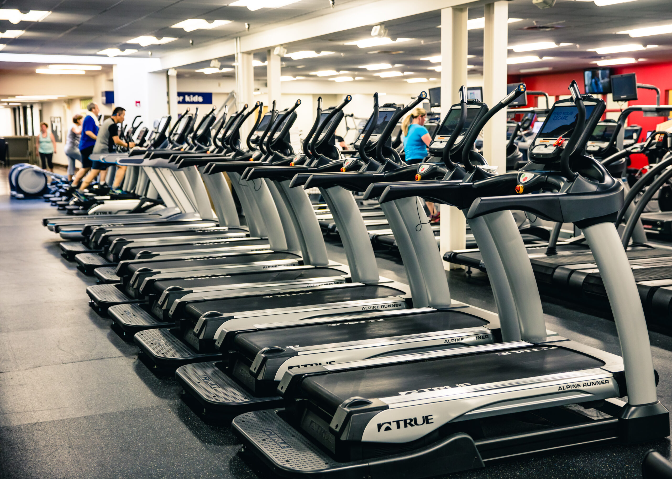 A row of treadmills in a gym, with several people using the machines. The gym has bright lighting and additional exercise equipment visible in the background.
