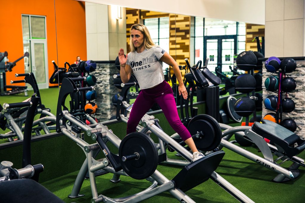 A woman wearing a grey OneLife Fitness shirt and purple leggings works out on a leg press machine in a gym with green turf flooring, mirrors, and various exercise equipment visible in the background.