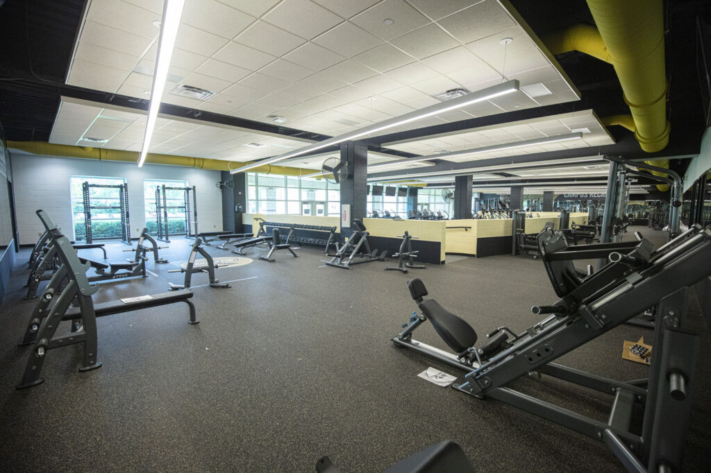 A spacious gym with weightlifting equipment, benches, and machines on rubber flooring. Large windows provide natural light, and mirrors line the walls. Yellow ductwork and a light-colored ceiling are visible. The gym is empty.