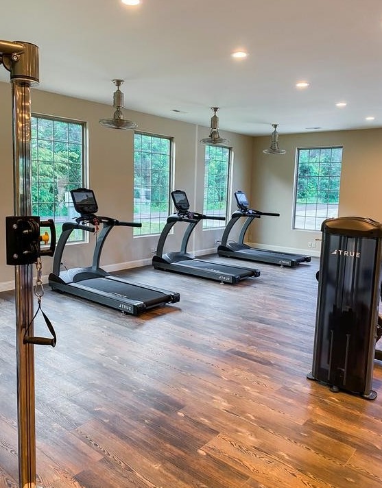 A modern gym with wooden floors, large windows, two treadmills, and exercise equipment. The room is well-lit with ceiling lights and has a view of greenery outside.