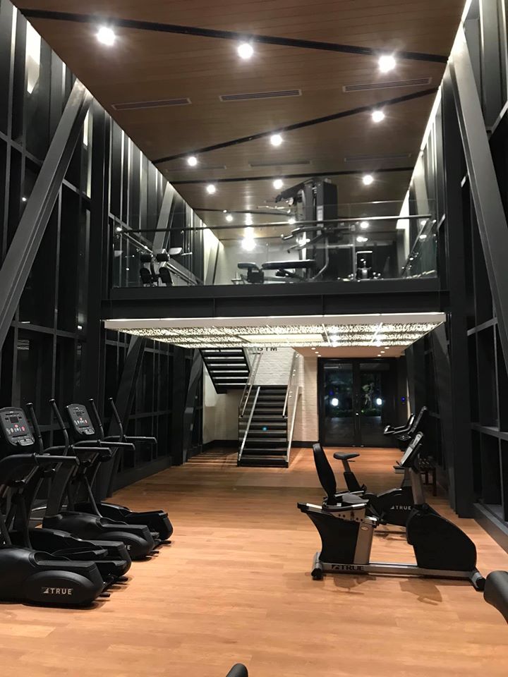 A modern, well-lit gym with large windows, treadmills, stationary bikes, and strength training equipment. A staircase leads to an upper mezzanine level with additional machines. The floor is wooden.