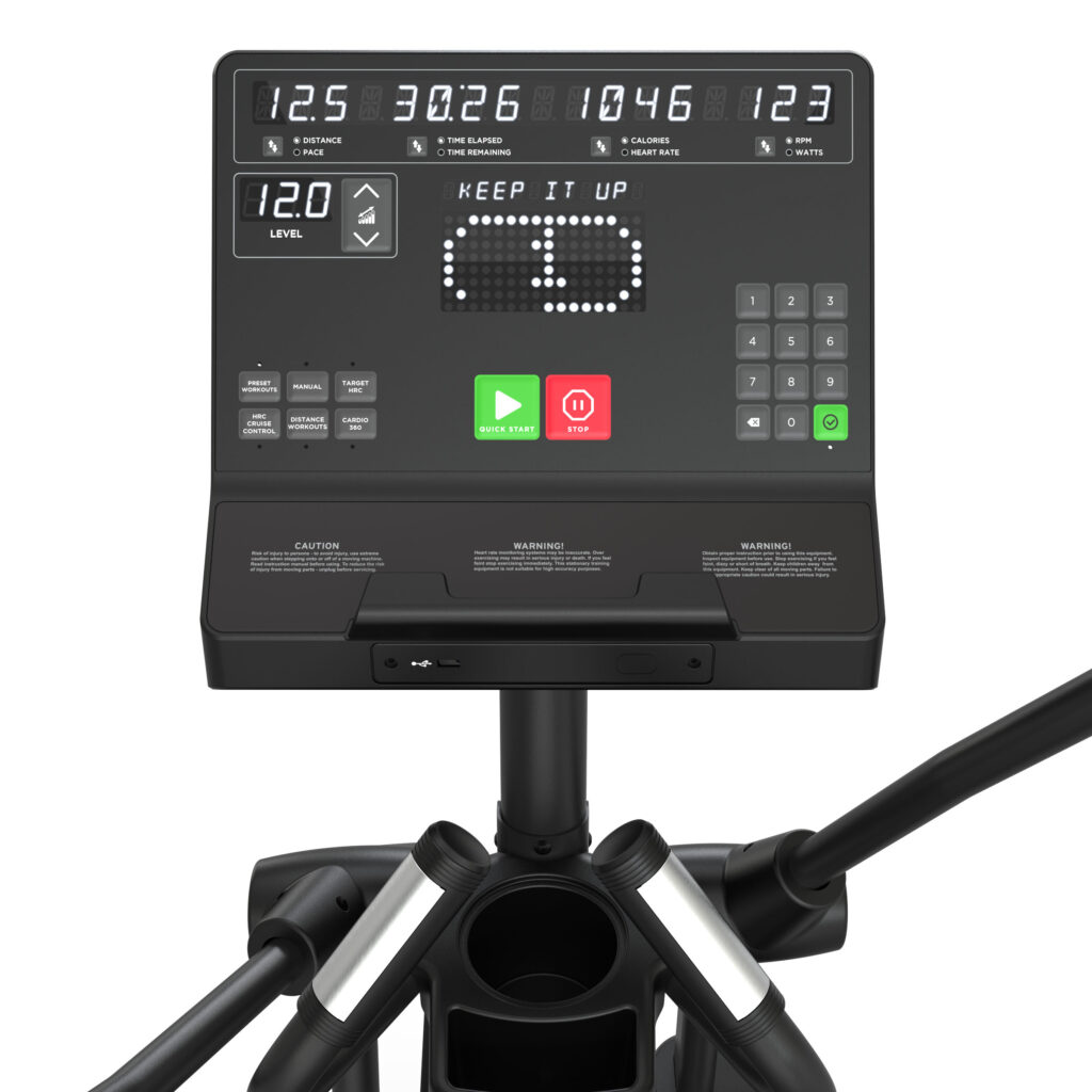 A close-up view of a black exercise machine console displaying workout metrics and a pixelated KEEP IT UP message, with various buttons, a number pad, and handles visible below the screen.