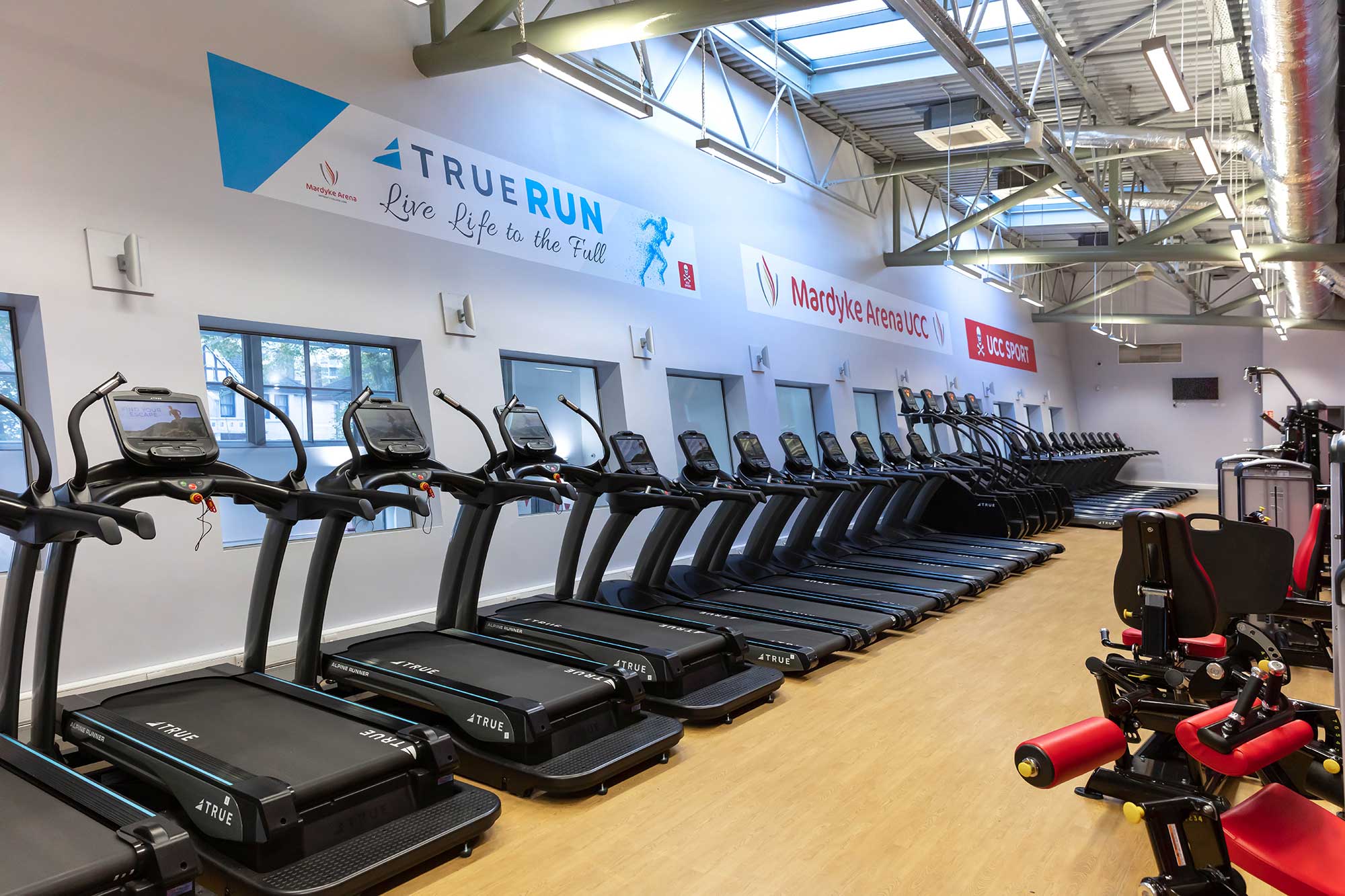 A modern gym with rows of treadmills lined up against a wall of windows, bright lighting, banners on the wall, and some strength-training equipment visible in the foreground.