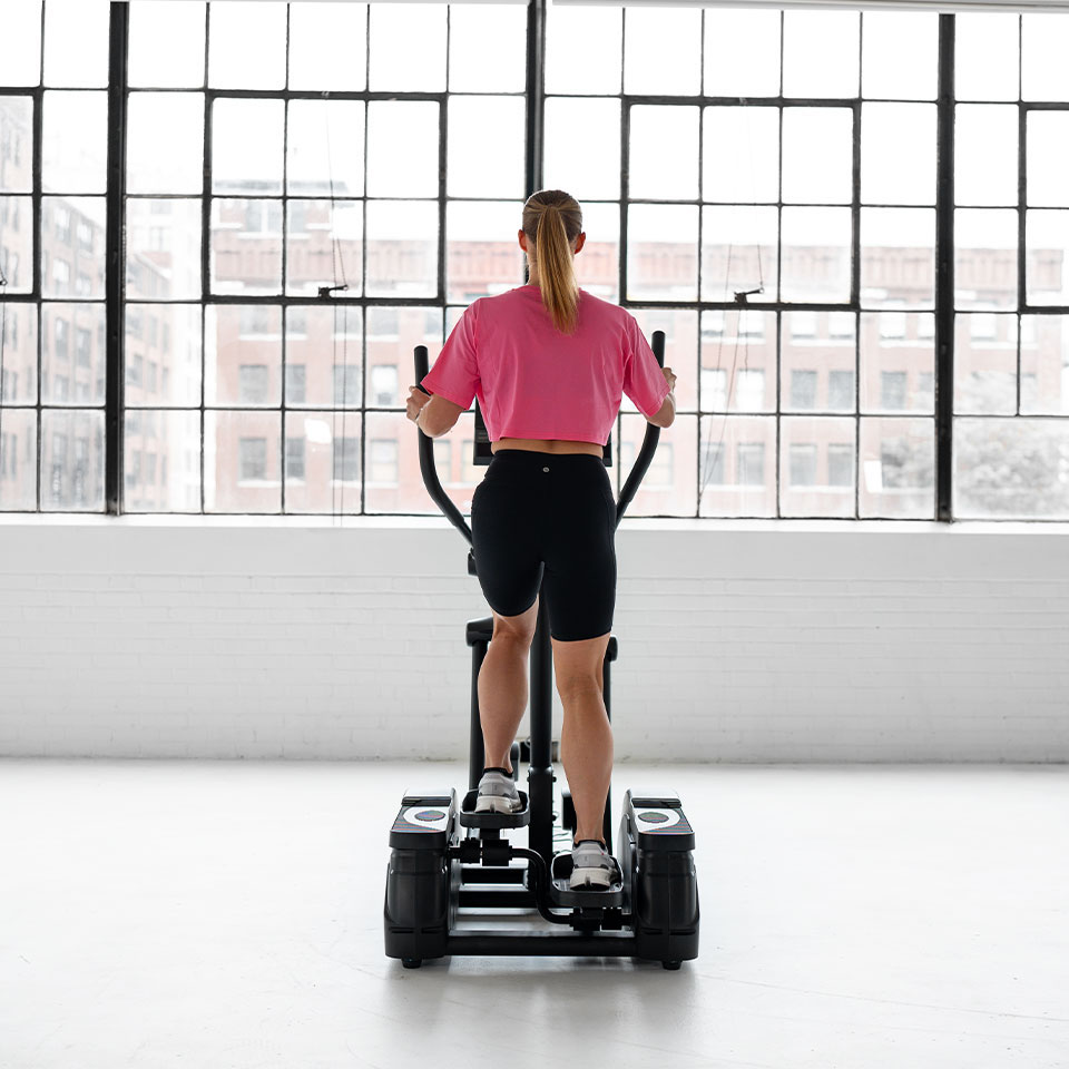 A woman wearing a pink shirt and black shorts exercises on an elliptical machine in a bright, spacious gym with large industrial-style windows overlooking city buildings.