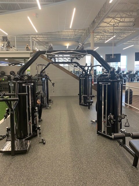A modern gym with multiple black cable exercise machines arranged in two rows facing each other, gray carpeted floor, and large windows letting in natural light. A staircase and mezzanine are visible in the background.