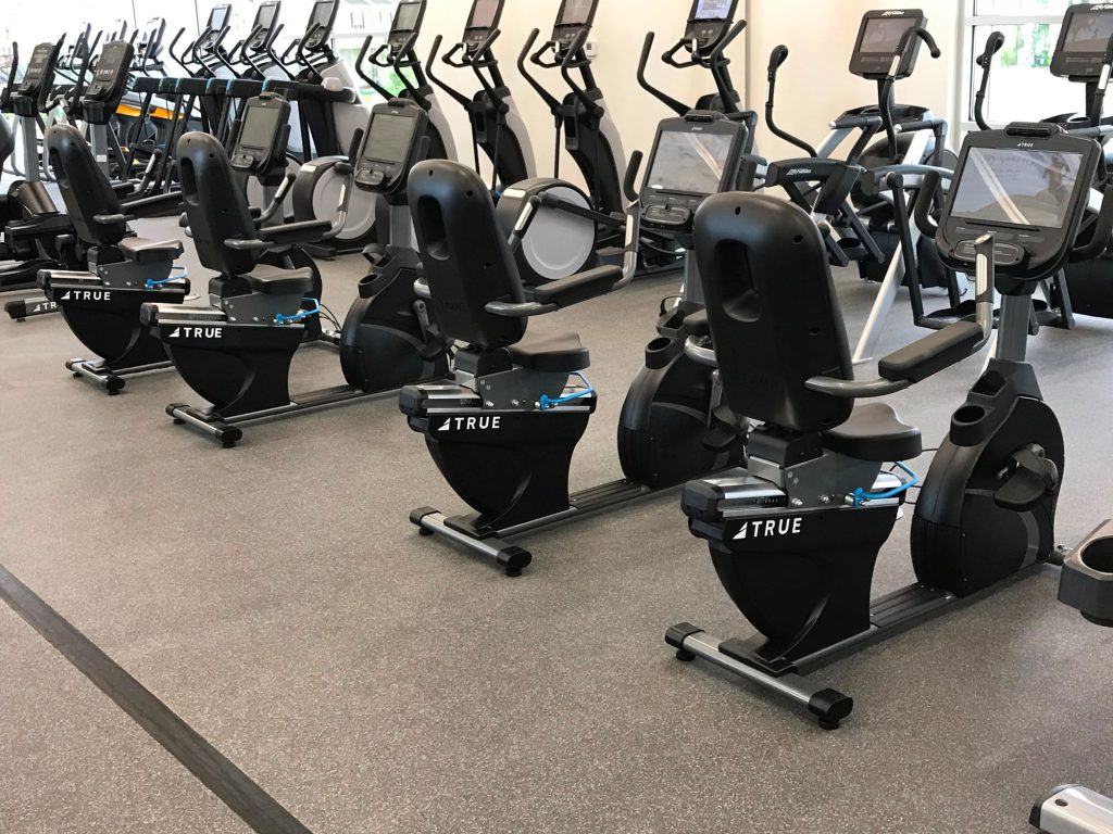 Rows of stationary recumbent exercise bikes and elliptical machines are neatly lined up in a gym with gray flooring, ready for use. The fitness equipment is modern and unoccupied.
