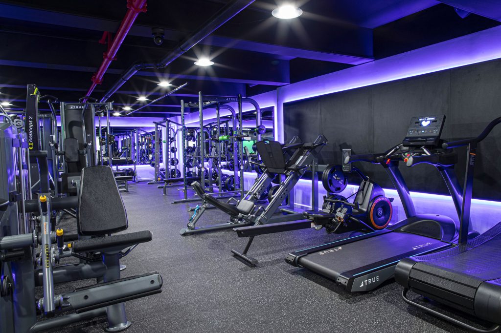 Modern gym with various exercise machines, including treadmills, leg press, and weightlifting equipment. The room features black walls, purple LED lighting, and a clean, organized layout. No people are present.